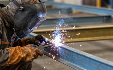 A close-up action shot of a skilled welder in a North American workshop, wearing protective leather gear, fabricating a custom steel deck railing. Bright sparks contrast with the steel blue and brass metal background.