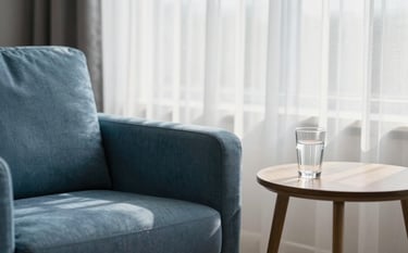 A close-up photograph of a bright, modern consultation room in North America. The scene features a comfortable soft blue fabric armchair next to a small wooden table with a glass of water, soft morning light filtering through thin white curtains, creating a calm and empathetic atmosphere.
