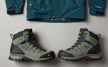 A flat-lay photograph of professional hiking boots and technical outdoor apparel. Clean composition on a neutral background, featuring deep teal and sage green accents. Sharp, high-contrast lighting to show durable textures. Global / Western aesthetic.
