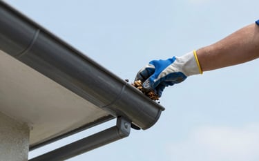 Close up of a clean, well-maintained roof gutter system with a professional hand in a glove removing debris. Bright sky background, conveying a sense of preventative care and clarity. Brand colors #1A2F40 and #347BBF subtly integrated.