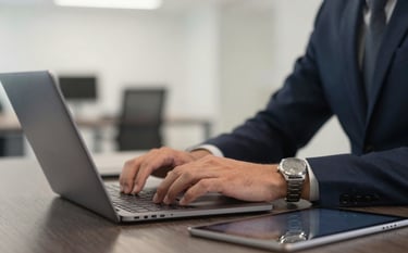 A close-up of a professional in a North American / US office setting using a laptop and a tablet. A muted silver watch is visible on the wrist. The desk is a dark charcoal wood, and the background is a blurred, efficient open-plan office with soft off-white accents.