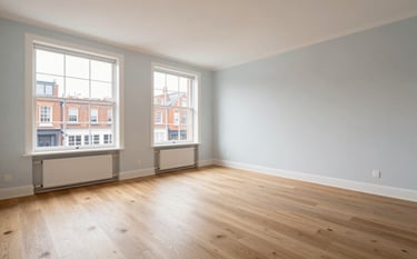 A wide-shot photograph of an empty, spotless British apartment living room with light oak flooring and pale blue-white walls. Large windows overlook a classic brick street. The lighting is soft and even, emphasizing a refreshed and ready-to-move-in feeling of excellence.