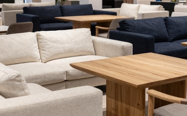 A high-end showroom in a Western European / Dutch commercial district featuring a set of brand new modern sofas and oak dining tables. The lighting is bright and natural, highlighting textures. The interior uses a palette of soft off-white and deep midnight navy accents.