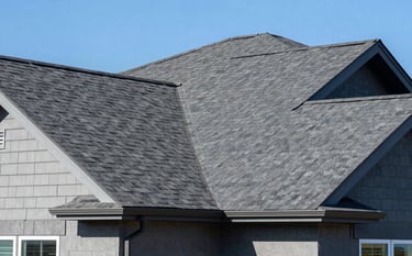 A close-up photograph of a modern North American / US suburban home featuring high-quality charcoal gray asphalt shingles. The lighting is bright and clear, highlighting the clean lines and professional installation of the roof. The brand's steel blue and soft blue tones are reflected in the clear sky and house accents.