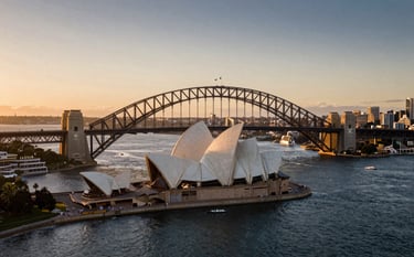 An aerial cinematic shot of the Sydney Harbour Bridge and Opera House at sunset, soft golden and dark blue tones, representing the prestigious and desirable Australian migration destination, high-end travel photography style.