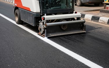 A professional wide-angle shot of a road marking machine applying a crisp, white thermoplastic line on a black asphalt road in a South Asian Indian urban environment. Bright daylight with high contrast and sharp focus on the machinery and the line.