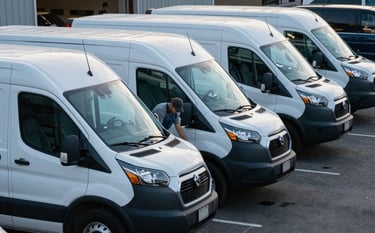 A row of white North American / US commercial delivery vans parked in a lot. A Momentum Glass Repair mobile service van is visible, with a technician working on one of the vehicles. Professional, reliable, and corporate mood. Soft lighting, Steel Blue and Dark Navy tones.