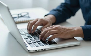 Close-up of professional hands typing on a laptop with a high-end metal finish. On the desk, there is a pair of spectacles and a notebook. The lighting is bright and natural, reflecting efficiency. Colors are tuned to the brand mood using #0A1C29 and #EAF2F4 tones.