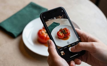 A close-up, high-angle shot in a cozy North American bistro where a hand is using a smartphone to capture a beautifully plated dish featuring deep ripe crimson heirloom tomatoes. The lighting is soft and natural, emphasizing the artisanal textures of the food and the matte forest green napkins on a crisp parchment-colored tablecloth.