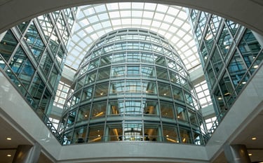 Low angle photography of a modern, glass-domed North American government building atrium. The scene is bright and airy, reflecting innovation and transparency with light blue and grey tones throughout the architecture.