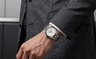 A man in a charcoal grey tailored suit jacket, focusing on a silver luxury watch on his wrist, minimal masculine environment with a dark leather and steel background.
