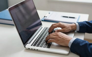 A close-up of a professional's hands typing on a high-end laptop in a bright North American / US home office. Beside the laptop is a neatly organized folder and a planner. The color palette features deep navy blue and pale frost blue for a trustworthy feel.