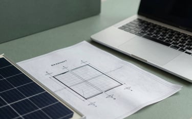 Close-up, high-angle professional photograph of a workspace. Modern blueprints are spread out next to a laptop and a piece of solar technology. Muted sage green and dark slate green office accents create a sophisticated, forward-thinking mood.