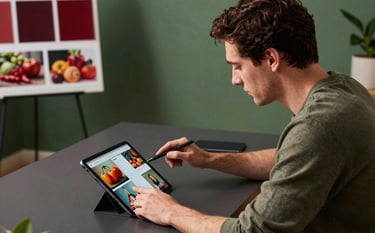 A professional in a modern North American / US creative studio environment sitting at a dark grey desk, planning a social media campaign on a sleek tablet. The room features matte forest green accents and warm, natural lighting. In the background, a mood board shows deep ripe crimson color swatches and photos of fresh produce. High-angle, professional photography.