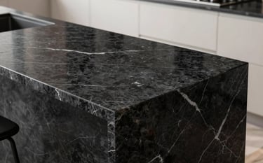 A high-end modern kitchen featuring a thick, polished deep charcoal black marble countertop with elegant white veining. The scene is lit by soft natural light hitting the reflective surface, highlighting the smooth texture and the robust quality of the stone. Background shows soft off-white cabinetry.