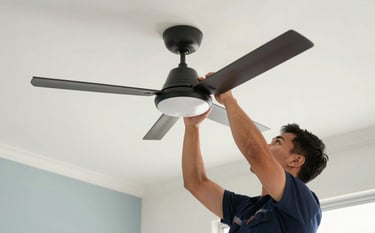 A close-up photograph of a professional technician in a modern Southeast Asian / Malaysian home, installing a sleek ceiling fan. The room is bright and clean with walls in soft off-white and pale blue-grey accents. The focus is on precision and safety, using professional tools. Natural lighting.