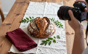 A professional photographer shooting a flat-lay of artisanal sourdough bread and fresh herbs on a rustic wooden table. The scene is bathed in soft, natural Scandinavian-style light. Deep Ripe Crimson napkins and Matte Forest Green garnishes provide color accents against the Crisp Parchment table runner.