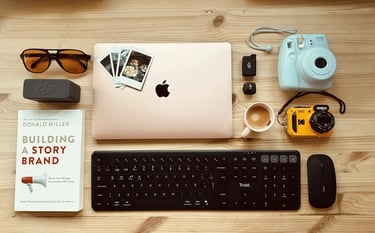Flat lay of a workspace with a MacBook laptop, Instax camera, Trust keyboard, and Building a Storybrand book.