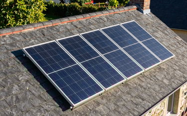 A high-angle photograph of high-efficiency solar panels installed on a traditional Central European / French slate roof. The scene is bathed in bright afternoon sunlight, reflecting a clean and sustainable energy vibe. The background shows a hint of a lush green garden in vibrant leaf green.