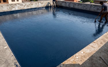 A wide angle shot of a South Asian / Indian building terrace during the day. Workers are applying a thick, protective layer of dark blue waterproofing material across the concrete floor to prevent leaks.