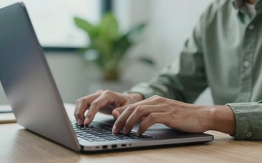 A close-up photograph of a professional developer's hands working on a sleek laptop in a bright office environment. Accents of muted sage green in the office plants and deep steel blue on the laptop cover.