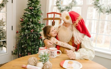 A smiling young girl hugging Santa Claus at a holiday table with cookies, cocoa, and a Christmas tree.