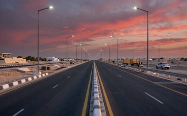 A professional wide-shot photograph of a road infrastructure project in a Middle Eastern / Saudi Arabian urban setting. It features a newly paved asphalt highway with modern lighting systems. The scene is shot during the golden hour, with hints of rich crimson in the sunset sky.