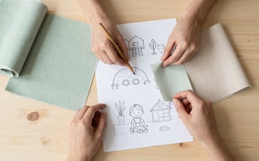 Professional top-down shot of a minimalist wooden workspace. Hands are seen arranging child-friendly design sketches and swatches of soft sage green and beige fabrics. The aesthetic is clean, modern, and highly professional.