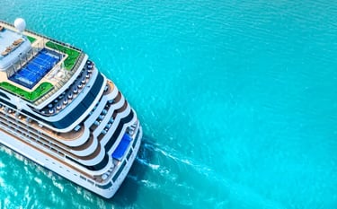 Aerial view of a luxury cruise ship sailing through turquoise ocean water with sports courts and sun decks visible.