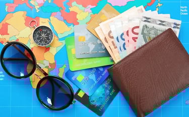 A brown leather wallet with Euro bills, credit cards, sunglasses, and a compass on a colorful world map.