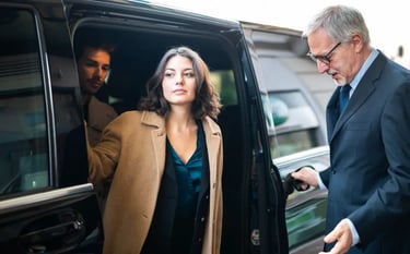 Professional chauffeur holding a car door open for a businesswoman exiting a luxury black SUV.