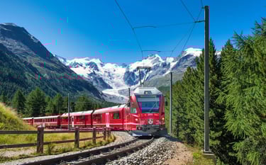 A red Bernina Express train traveling through the Swiss Alps with snow-capped mountains and green pine trees.