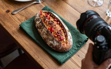A top-down food photography scene in a European / French bistro. A rustic wooden table with artisanal bread and dark forest green napkins. A professional camera is visible in the corner of the frame, capturing the deep red tones of fresh ingredients.