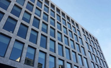 A wide-angle shot of a sleek, contemporary corporate building with large glass windows reflecting a clear blue sky. The architecture is sophisticated and premium. Global / Spanish-speaking setting. Lighting is crisp and bright. Palette incorporates light grey-blue and white.