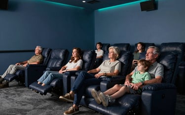 A spacious North American / US home cinema room with several comfortable dark navy reclining chairs. Multi-generational family members are gathered. The walls are muted blue, and the lighting is dim with soft teal LED accents.