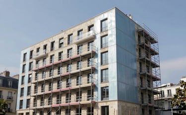 Wide shot of a contemporary apartment building in a European / French city during a renovation. Scaffolding is visible, and part of the facade features new light steel blue cladding and beige stone finish under a clear sky.