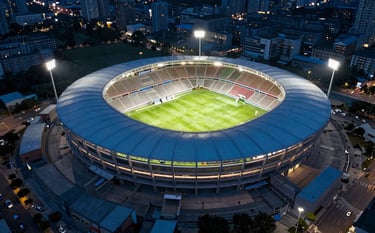 A high-altitude aerial photograph of a massive outdoor soccer stadium at night, brilliantly lit by Big Dipper lighting systems. The surrounding urban landscape is dimly lit in steel blue and dark blue, emphasizing the vibrant, clear light of the venue.
