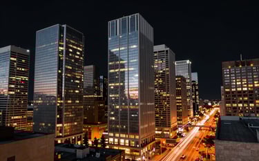 A panoramic twilight view of a North American / US metropolitan financial district. The sleek glass skyscrapers reflect a deep black sky, with vibrant orange light trails from traffic creating a sense of high-speed global connectivity.