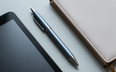 A top-down, clean macro photograph of professional tools on a pale grey surface: a high-quality pen, a digital tablet, and a clean leather planner in a North American / International workspace. Colors are muted steel blue and soft silver.