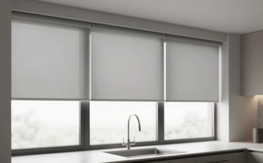 A modern, clean kitchen with large windows fitted with sleek motorized solar shades. The shades are a light gray #DEDBD6 tone, partially lowered to show the view while reducing glare. The setting is bright and professional, emphasizing a smart home aesthetic.