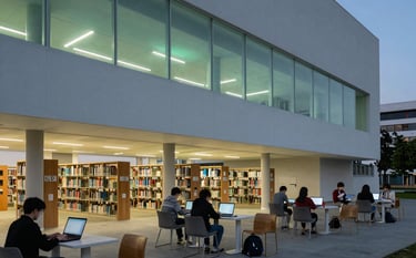 An architectural photography shot of a sleek, minimalist university library interior at dusk. Students are working on thin laptops under soft green LED ambient lighting. Global / English-speaking.