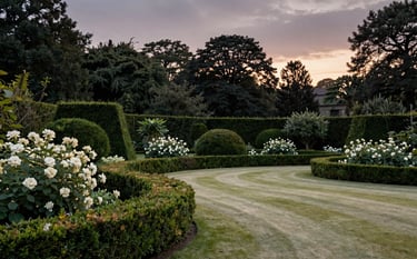 A wide landscape shot of a manicured estate garden at twilight, with soft off-white blooms, dark forest green sculpted hedges, and a serene pale olive lawn.