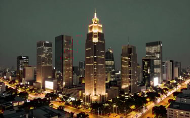 A stunning evening shot of the Gurugram Cyber City skyline. The towers glow with warm light against a dark slate green twilight sky. Below, the urban infrastructure reflects a modern South Asian / Indian commercial hub with flowing traffic and sleek, sophisticated architecture in a professional setting.