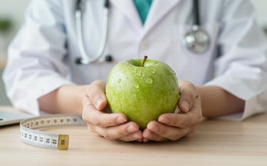A close-up of a nutritionist's hands holding a green apple and a measurement tape on a clean, light wood desk. The scene is sophisticated and trustworthy, with a soft blur in the background suggesting a professional clinic environment using colors #52796F and #F5F5DC.
