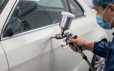 Close-up of a professional automotive paint job in a Canadian spray booth. A technician wearing protective gear uses a high-precision spray gun on a metallic car door. The environment is spotless and reflects a palette of off-white and dark navy.