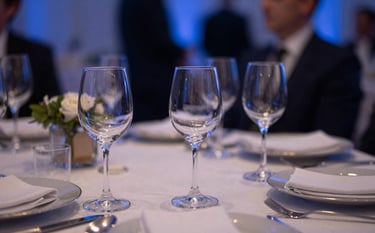 A sophisticated evening corporate gala event in a North American setting. The shot is a close-up on elegant table settings with glass details reflecting muted steel blue and dark navy blue ambient light, capturing a sense of reliability and clarity.