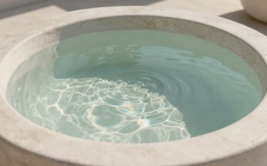 A close-up photograph of clear, serene water rippling gently in a stone vessel, with soft morning light filtering through. The scene is set in a calm, modern facility in Redmond, Oregon, North American / US. The color palette features soft off-white and hints of muted green.