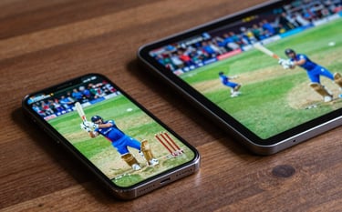 Close-up photography of a modern smartphone and tablet placed on a wooden table in a Global / Indian-influenced setting. The screens show a high-energy live cricket match with vibrant colors, emphasizing clarity and portable technology.
