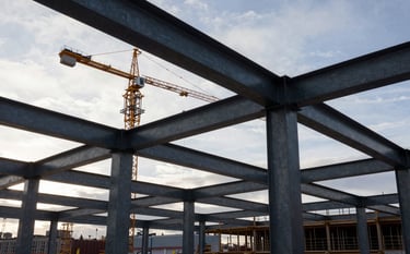 Professional architectural photography of a large-scale construction site in a Northern European / Finnish urban environment. Focus on steel structural beams, a crane silhouette against a soft cloud white sky, industrial slate grey textures, and high-contrast lighting with deep charcoal black accents.