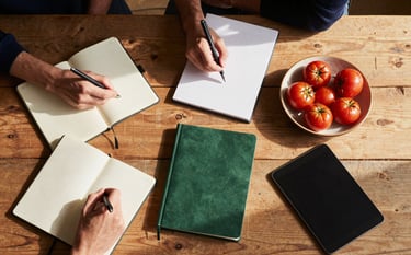 Top-down view of a rustic wooden table where a marketing team is planning a campaign. There are notebooks, a tablet, and a ceramic bowl of small red tomatoes. Warm natural sunlight filters in, highlighting Matte Forest Green and Deep Ripe Crimson accents. Scandinavian minimal style.